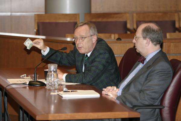 Two older men sitting at a court room table. One is holding up a dollar bill