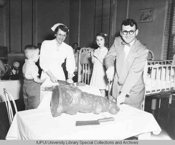 Man in a suit and bowtie and a woman in a nurse uniform carve a large chocolate bunny while being flanked by children