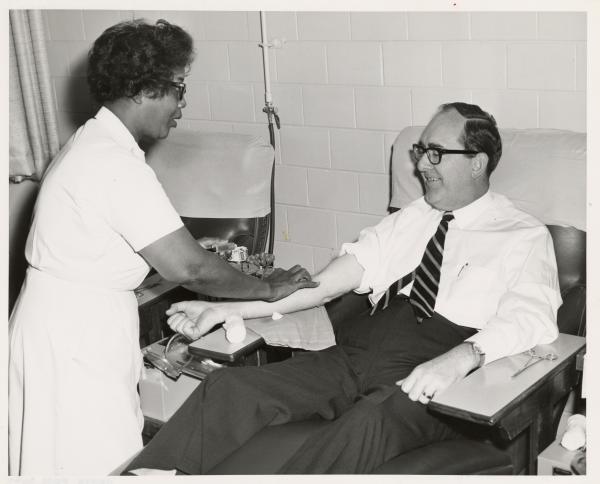 Older man in a tie seated while a nurse draws his blood