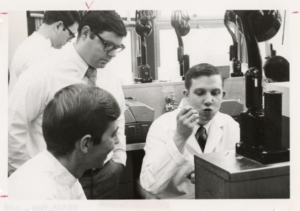 A young man in a lab coat appears to be blowing on a metal utensil. Two other men watch