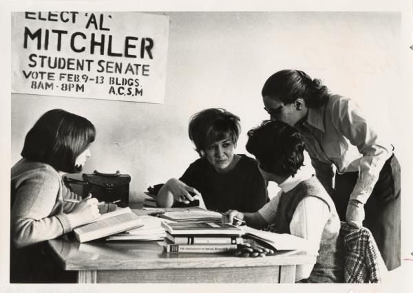 Four young women huddled around a table. Above them is a poster that read Elect Al Mitchler Student Senate Vote Feb 9-13 Bldgs 8AM-8PM ACSM