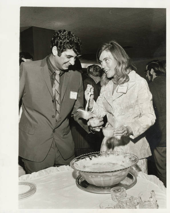 Balck and white photograph. A young white woman is ladeling punch into a small cup help by a man in a suit with curly dark hair. The man is laughing and she is smiling. She also holds a small cup. People can be seen in the backgroundm as if at a party.