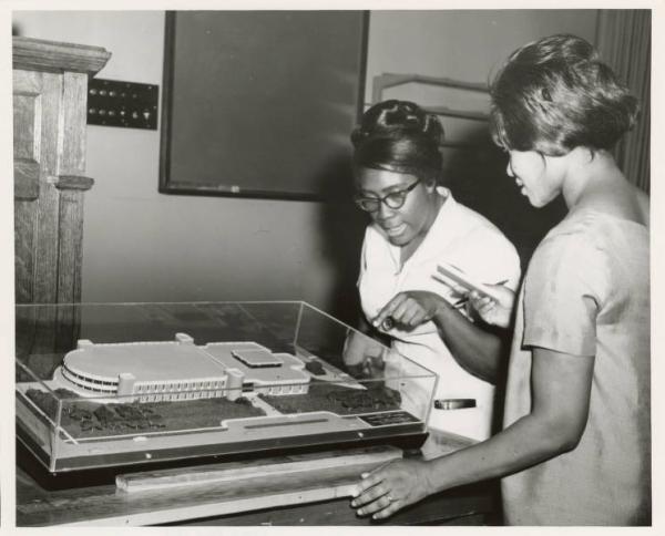 Black and white photograph. Two young black women are looking at a miniature diorama of a large displayed under a clear container. One of the women is wearing glasses and has a beehive style hair-do. She is pointing to the model and talking. The other woman is holding a notepad and pen in her right hand. In the background we can see a blackboard, wooden podium, and several light switches.  