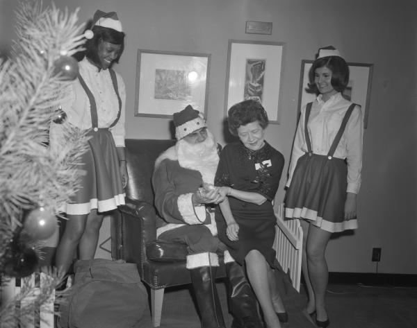 Black and white photograph. An older white woman with an updo and a dark blouse and skirt is seated on a man dressed like Santa Clause with a fake beard. He is holding her hand and has a prop bag on his righthand side. On either side of them stand two young women, one white and one black, dressed like elves. In the foreground is a decorated Christmas tree. In the background we can see three framed paintings and an electrical socket.