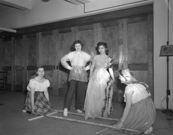 Black and white photograph. Two young women are standing, each with their right foot in front, between parallel bamboo stalks. Two other young women are squatting on either side, holding the bamboo stalks. One of the standing women is wearing an ornate dress and is of asian descent. The other standing woman has dark short hair, and is dressed in slacks and a white button up shirt. Over the top is a gauzy shirt of some kind. The two kneeling women are in white peasant shirts and skirts with plaid.