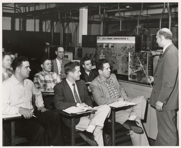 Black and white photograph. A class of young white men, watch intently as an older, balding white man stands up front teaching. The teacher has a thin pointer in his hand and is using it to point at a machine that is open and filled with cables. Another older man, wearing a suit and tie, is sitting with the students, smiling broadly. On the table, next to the open machine is another machine that is labelled RCA Dynamic Demonstrator 2. The background looks somewhat industrial.