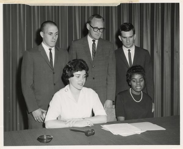 Black and white photograph. Three white men in suits stands behind two seated women. On the table in front of them is judge's gavel and several sheets of paper. Everyone is looking at the spread papers with concerned expressions on their faces. The leftmost man has a shaved hair style and the one in the middle wears big glasses. The leftmost woman is white with dark hair and is wearing a bright white blouse. The other woman is black, and is wearing a dark dress and a pearl necklace. Behind them appears to be a heavy curtain.