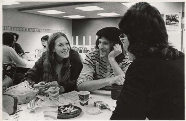 Black and white photograph. Three young people, one man and two women are in frame, seated around a table. One of the women is seated away from the camera, but we can still see that she is wearing thick glasses. The man has long dark hair underneath a black beret. He is resting his cheek on a hand and looking off camera smiling. The other women has long wavy hair and is wearing a hoodie and t-shirt. Between all of the students is an ashtray filled with cigarettes, several paper cups, some of which are scrunched up, and an open pack of cigarettes. In the background we can see another table of three students talking.