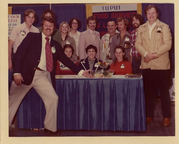 Color photograph. A man in a blue sports coat, tie and khaki pants stands in front of a table with his arm out, displaying it to the photographer. He is smiling and has a mustache and sideburns. On the table sits a clear fruit bowl, a magazine, and some other papers. It has a blue tablecloth with a white top. Behind the table sit three young women who are smiling. Each person has a round nametag affixed to their clothing. Behind them are nine other people, posing. A sign in the back reads, "IUPUI Food Services"