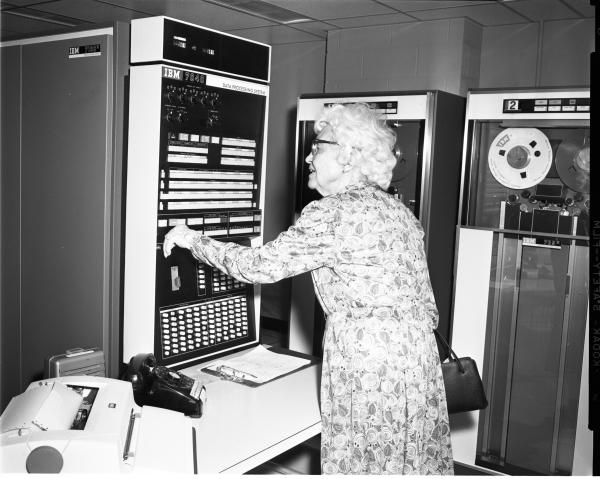 Black and white photograph. An older woman with white curly hair and glasses wearing a dress with a spiral pattern is facing away from the camera at a 45 degree angle. She is facing a computer from the 1960s, labeled “IBM 7040.” It is roughly 2 and ½ feet tall and 1 foot wide, with dials, switches, and buttons for the interface. Her left arm is raised, interacting with the machine. On the ame desk that the computer sits on is a printer, rotary telephone, and clipboard. In the background are other vintage data storage devices, with tape memory systems.