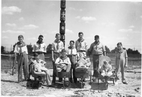 Black and white photograph. Four boys, aged seven to 11 sit in three wheeled wheel chair style devices, made of wood. Behind them stand seven other boys. All of them are wearing boy scout uniforms and are posing for the camera. Behind them all is a wooden stick fence and a totem pole carved with a sun and a fleur-de-lis. The ground is well worn dirt and the sky has puffy clouds. 