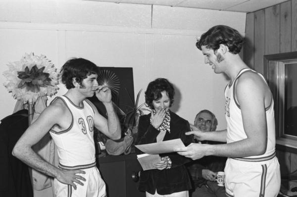 Black and white photograph. Two young men stand with an older woman. Both of the men are wearing basketball uniforms. The woman is laughing with a hand to her mouth while looking at a piece of paper that the taller of the two men is holding. He also has his hand on her shoulder. Behind them is an older man seated with a mug, smiling up at the woman. The taller man has the number 54 on his jersey, the other the number 30. In the background is a safe with a purse and plants on top of it and a giant flower made of tissue paper. One of the walls behind them is wood paneled. 