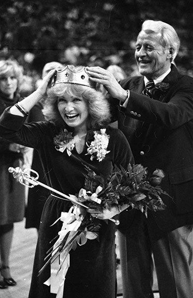 Old man places a crown on a smiling woman holding flowers and a scepter