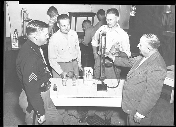 Four men, one in police uniform, around a table talking about a large glass tube connected to small beakers 