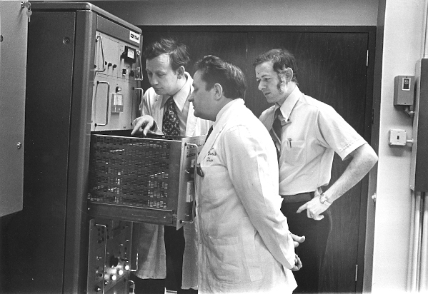 Three men in ties huddle around an open drawer of a large machine