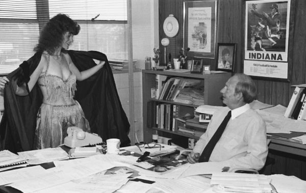 Man sitting in hir office looking at a woman who has opened her coat to reveal evening wear