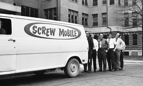 Black and white photograph. The year is 1971. A white van parked in front of a  multi-story campus building has a caption which reads Screw Mobile on its side. Many windows adorn the three stories viewable on the building. A large drainpipe snakes down the side of the building. There are leafless tree limbs showing on the right side of the picture. Lining up behind the van are Six smiling white men dressed in long sleeve shirts with ties.