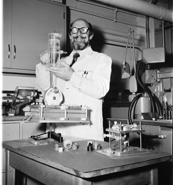 A man with a curled mustahce and baerd in a lab coat holds chemistry equipment over a table