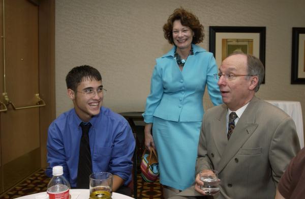 An old man in a suit and an old woman in a bright blue dress talk to a student in a tie