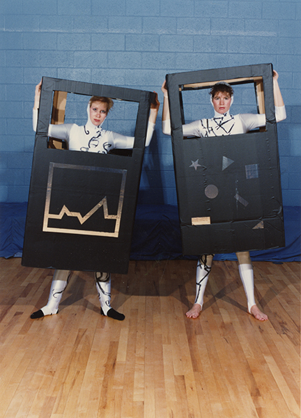 Two woman in shiny silver costumes hold cardboard boxes with space for their arms, and heads around their bodies, to resemble a calculator. The boxes are painted black and with geometric shapes on them.