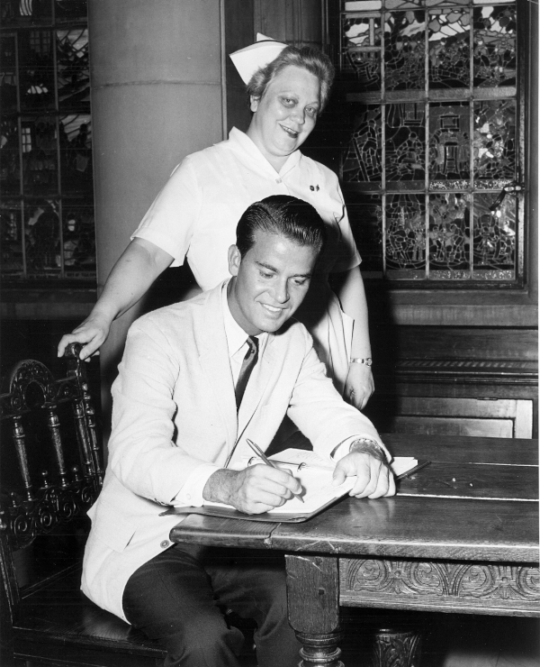 Dick Clark signs a guest book while a nurse behind him smiles into the camera