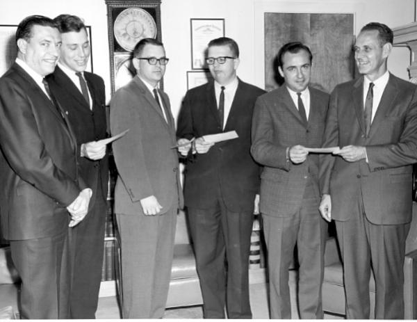 6 men in suits look into the camera holding checks