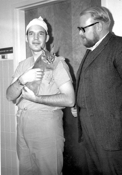 A man in hospital attire holds a baby lynx with a corpulant man in a suit looks at it