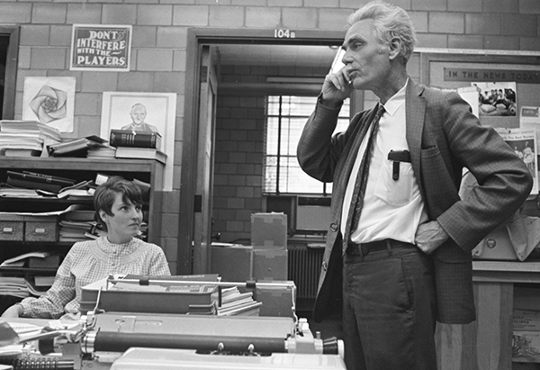 Older man in a suit stands facing a desk, his hand near his ear. A young woman looks up at him from the desk