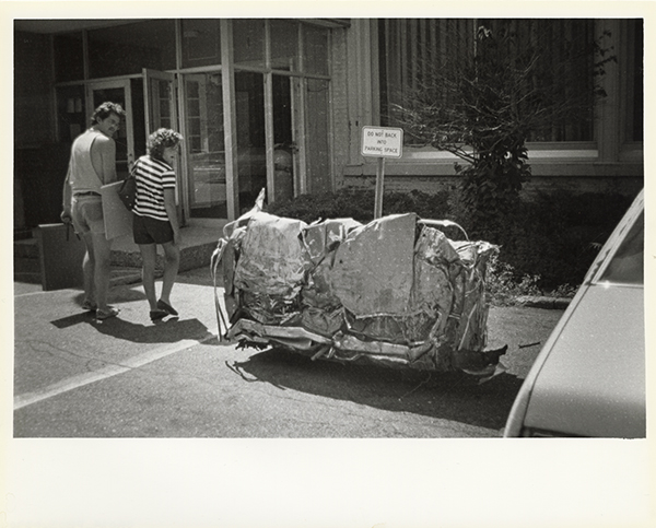 A car, crushed into a cuboid sits in a parking spot. Two students look at it over their shoulders