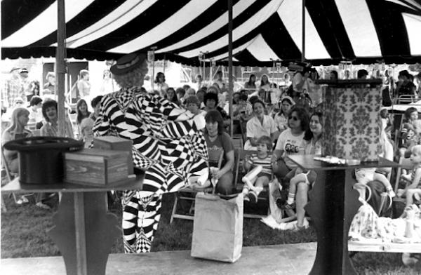 Man in a psychadelic black and white suit holds it open to a gathere crowd at a fair