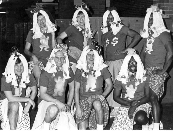 Eight men posing in skirts and wigs made out of mops