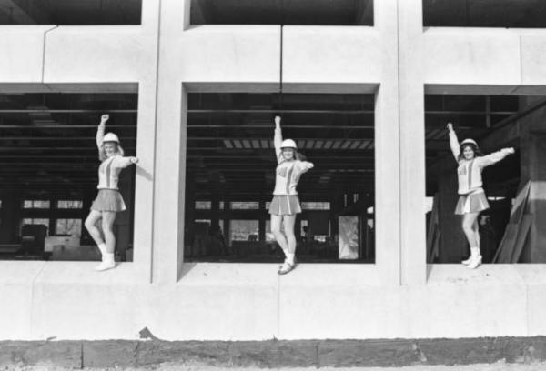 Three young women standing in a cheer pose between columns. They are wearing hard hats