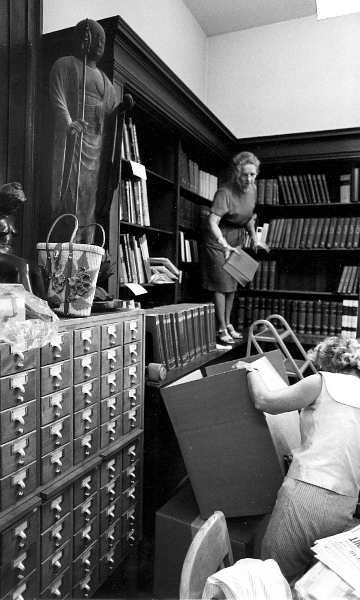 A woman standing on a desk throwing books at another woman holding a large box open