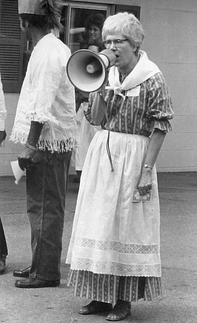 Old woman in pioneer style clothing speaks into a megaphone