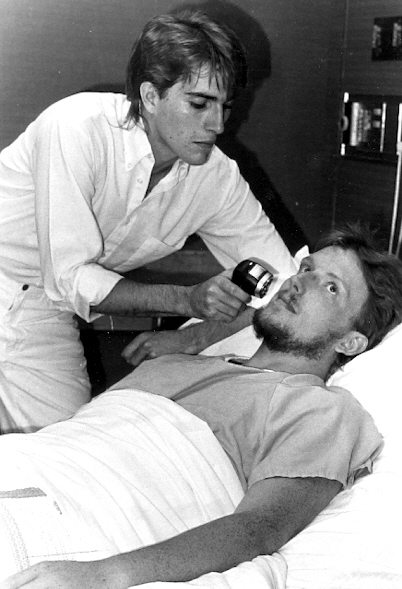 Bored looking young man lays on a hospital bed with another young man holding a testing device to his face