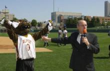 Older man with a microphone on a baseball field points to the IUPUI Jaguar mascot