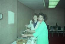 Two women using ladles to get a drink out of large bowls. One is smiling, the other has an expression of shock