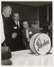 Two men and a woman look intently at a model of a drum. The drum has the words Worlds Largest Drum Purdue on it