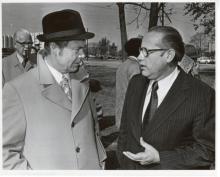 Two older man, one in a suit with slicked back hair, the other in a trilby and trenchcoat talk