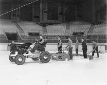 A young man on a tractor drives on an ice arena with a trailing plow. 5 others stand behind holding shop mops