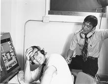 A young person seated in front of a machine with headphones on, laughs into the camera, while another student with a cigarette in his mouth seated behind him also laughs