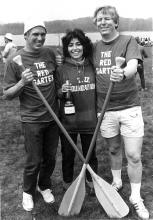 A young woman smiles holding a bottle of alcohol with two men flanking her, crossing oars in front of her.