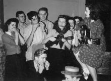 A mixed-gender band of musicians, 3 with harmonicas, one with a guitar, and one with a ukulele and a harmonica play for a small crowd.