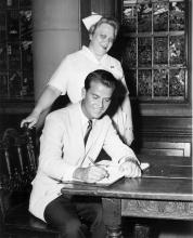 Dick Clark signs a guest book while a nurse behind him smiles into the camera