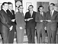 6 men in suits look into the camera holding checks