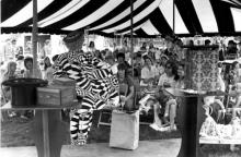 Man in a psychadelic black and white suit holds it open to a gathere crowd at a fair