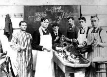 Black and white photograph. A medical school classroom from the early 20th century. Six young white men with serious expressions  stand in front of a chalkboard with notes drawn on it. One of the men has a handle bar mustache. They are surrounding a lab table with cadaver parts laid out on top. The men are wearing clothing consistent with the era, and they are wearing lab coats over the top. The man on the right is holding up a severed leg. 