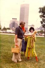 Woman in a clown outfit holds up a plush monkey that a man in a striped shirt pinches