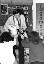 A man whose head is lowered, smiles up at the camera (giving him a sinister look), while handing items to two women reaching out for them