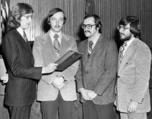 Four men stand around a plaque one of the men are holding. One of the men is looking directly into the camera